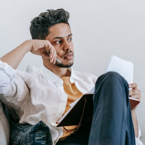Man in wit overhemd zit op een bank met een notitieboek in zijn handen en kijkt peinzend opzij.